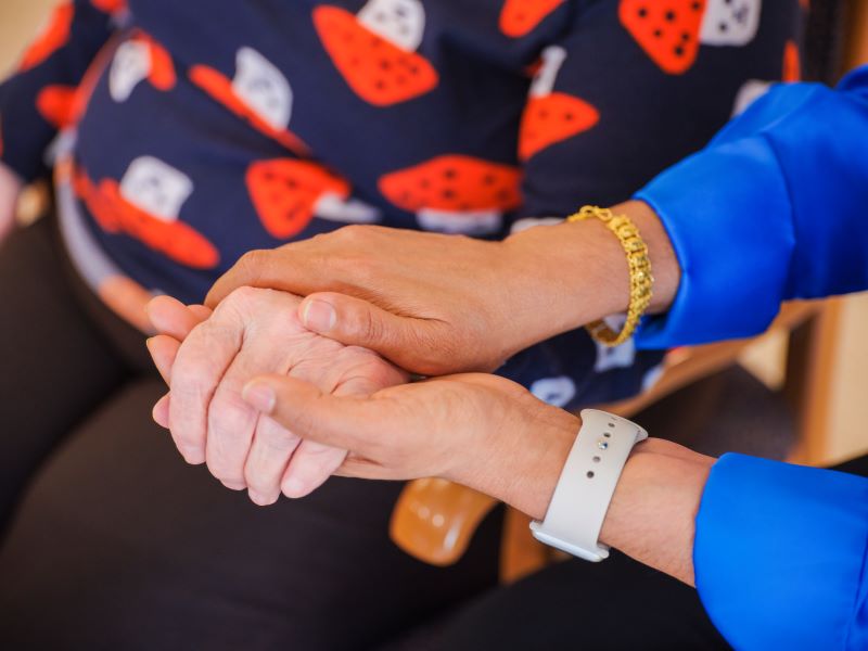 A person wearing a blue shirt holds and supports the hand of another person sitting down, who is wearing black pants and a patterned sweater.