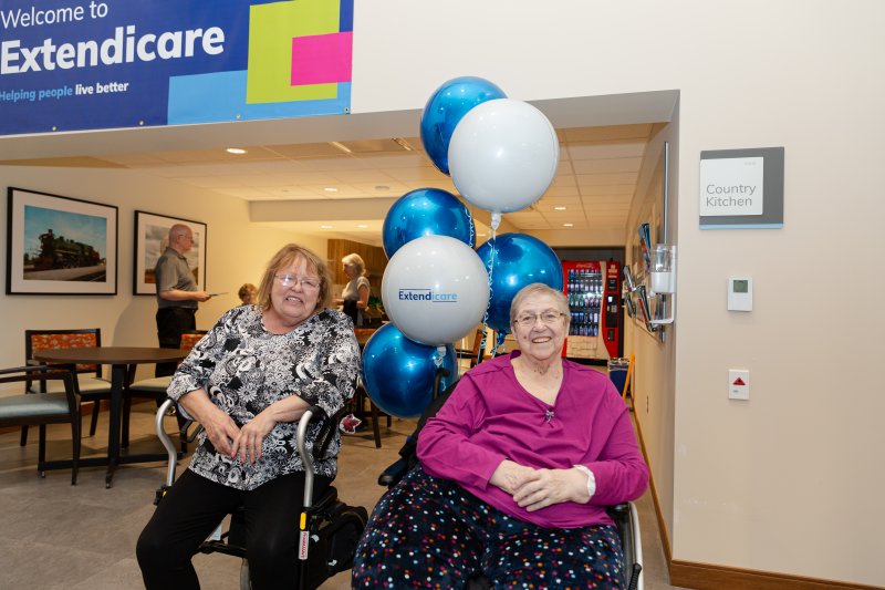 Two elderly women in wheelchairs smile under a cluster of Extendicare balloons in a care facility lobby near a sign labeled "Country Kitchen.