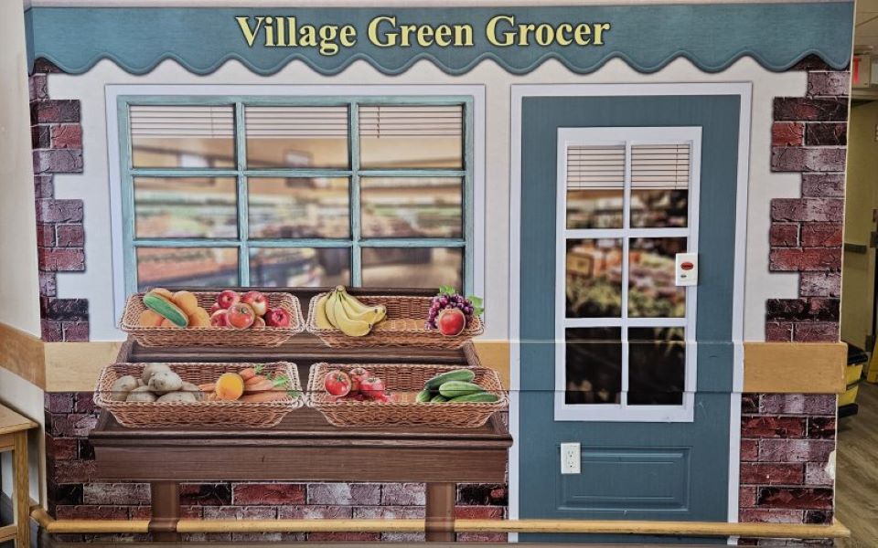 A mural of a storefront labeled "Village Green Grocer" shows baskets of produce including bananas, apples, potatoes, carrots, and cucumbers outside a door and window.