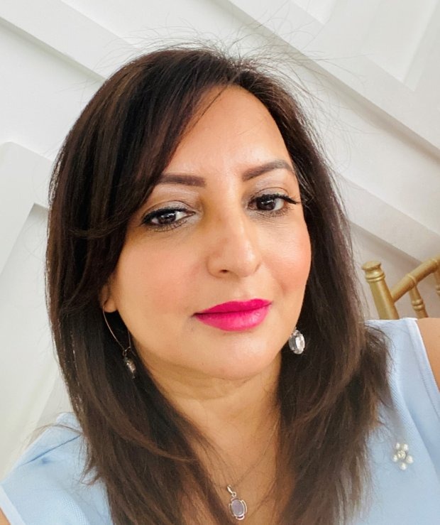 A woman with shoulder-length dark hair and bright pink lipstick wears a light blue top and dangling earrings, seated indoors.