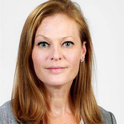 A woman with straight, shoulder-length blonde hair and light eyes, wearing a gray blazer and light top, poses against a plain white background.