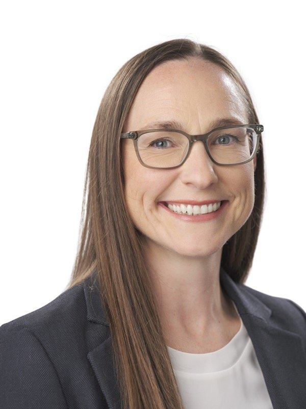 Woman with long straight brown hair, wearing glasses, a dark blazer, and a white top, smiling at the camera against a plain white background.