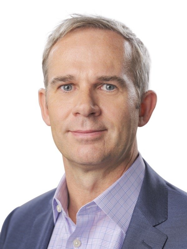 A middle-aged man with short blond hair wearing a blue blazer and a light purple checkered shirt, posing against a white background.