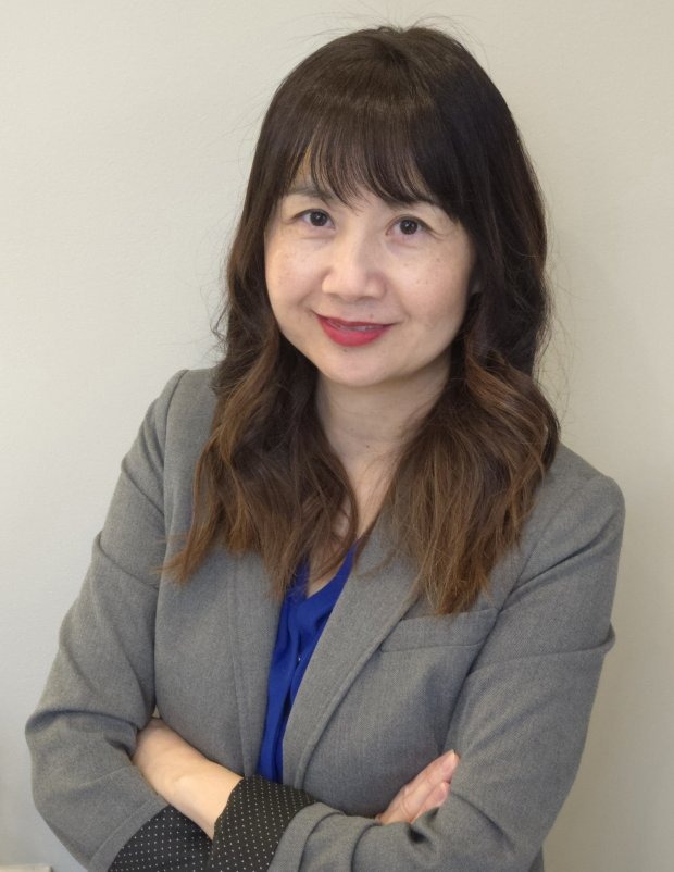 Woman with long brown hair wearing a gray blazer and blue top stands against a light-colored background with arms crossed, looking at the camera and smiling slightly.