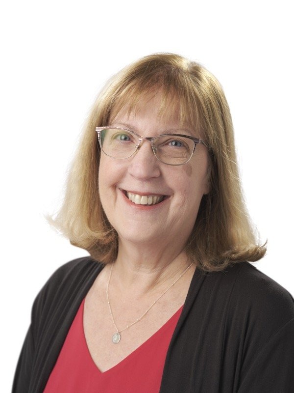 Smiling woman with shoulder-length light brown hair, wearing glasses, a red top, a black cardigan, and a necklace, poses in front of a white background.