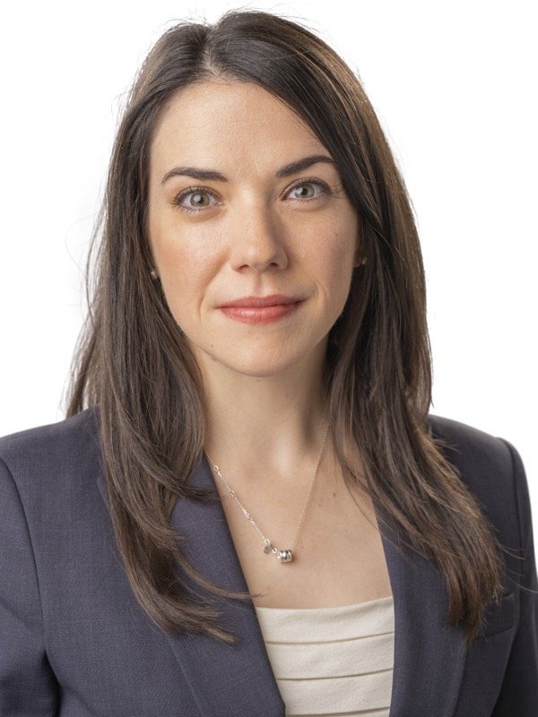 Woman with straight brown hair wearing a dark blazer and light top, facing forward against a plain white background.