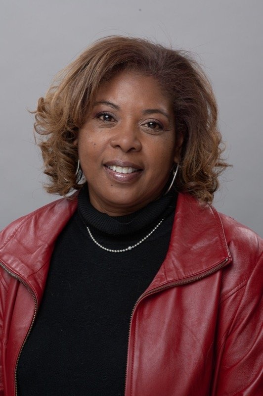 Woman with shoulder-length curly hair wearing a red leather jacket, black turtleneck, and pearl necklace, smiling at the camera against a gray background.