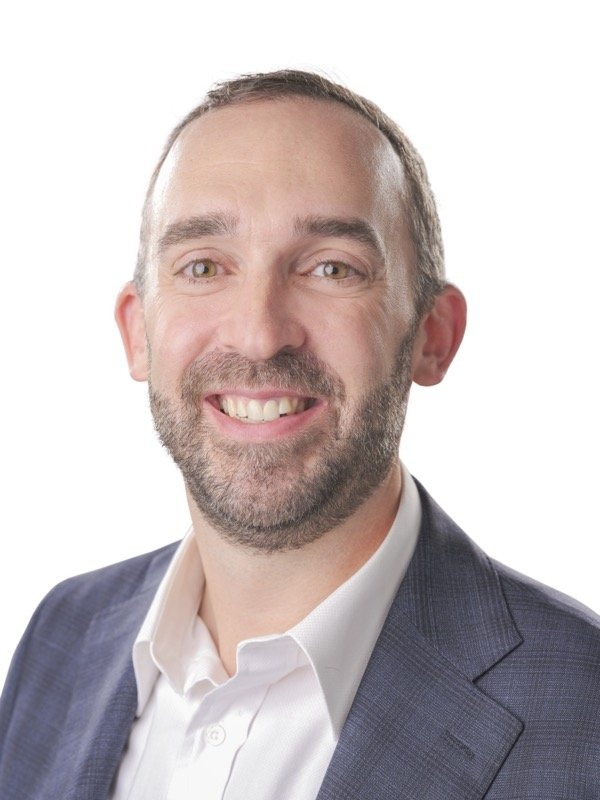 A man with short brown hair and a beard, wearing a white shirt and a blue blazer, smiling at the camera against a white background.