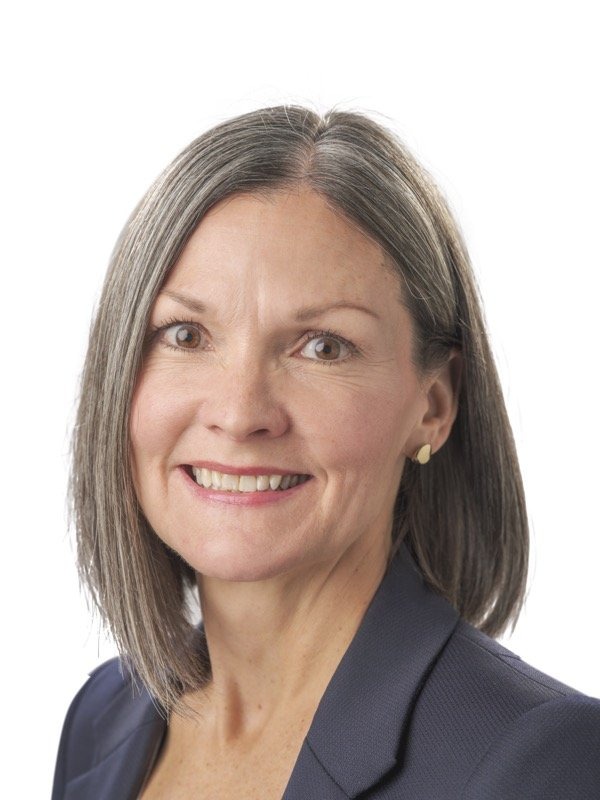 A woman with straight, shoulder-length gray hair, wearing a navy blazer and small earrings, smiles at the camera against a plain white background.