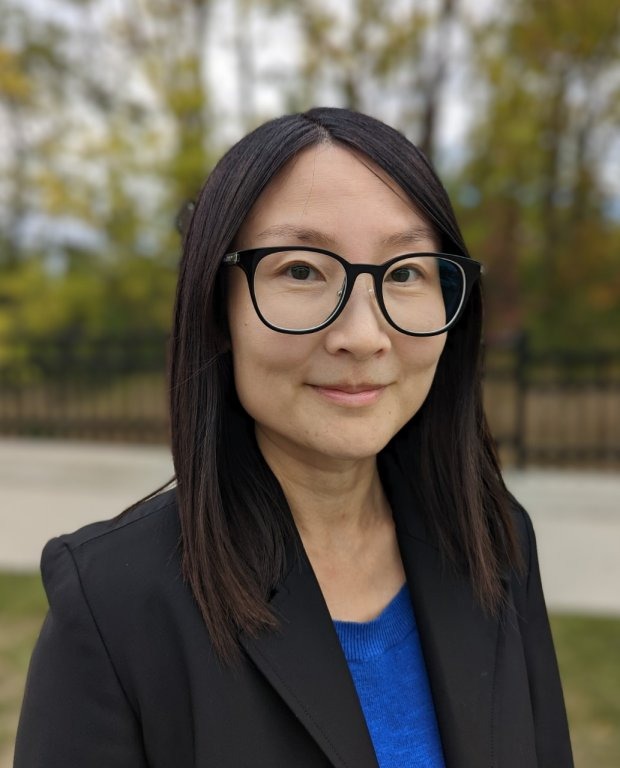 A person with straight dark hair and glasses wearing a black blazer and blue top stands outdoors in front of a blurred background with trees and a fence.