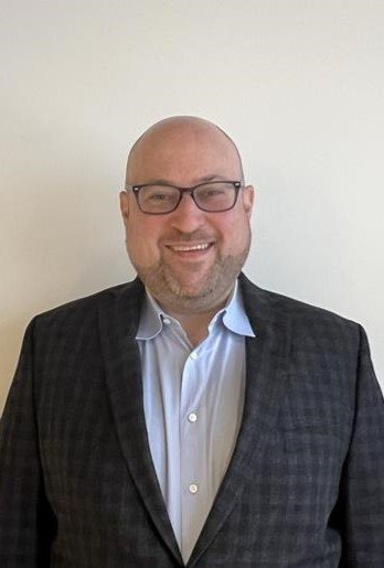 A man wearing glasses, a blue collared shirt, and a dark checked blazer stands smiling in front of a plain light-colored wall.