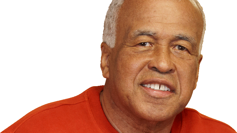 A man with short gray hair, wearing an orange sweater, is smiling at the camera against a plain background.