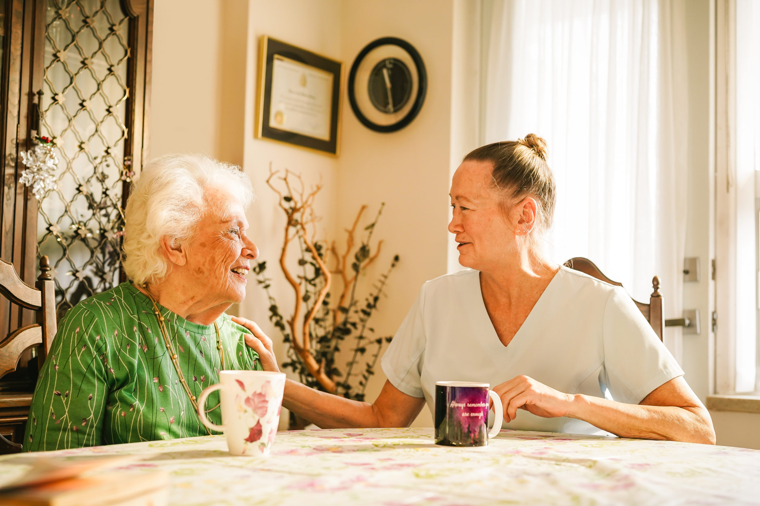 An elderly woman and a caregiver sit at a table with mugs, smiling and talking in a sunlit room.