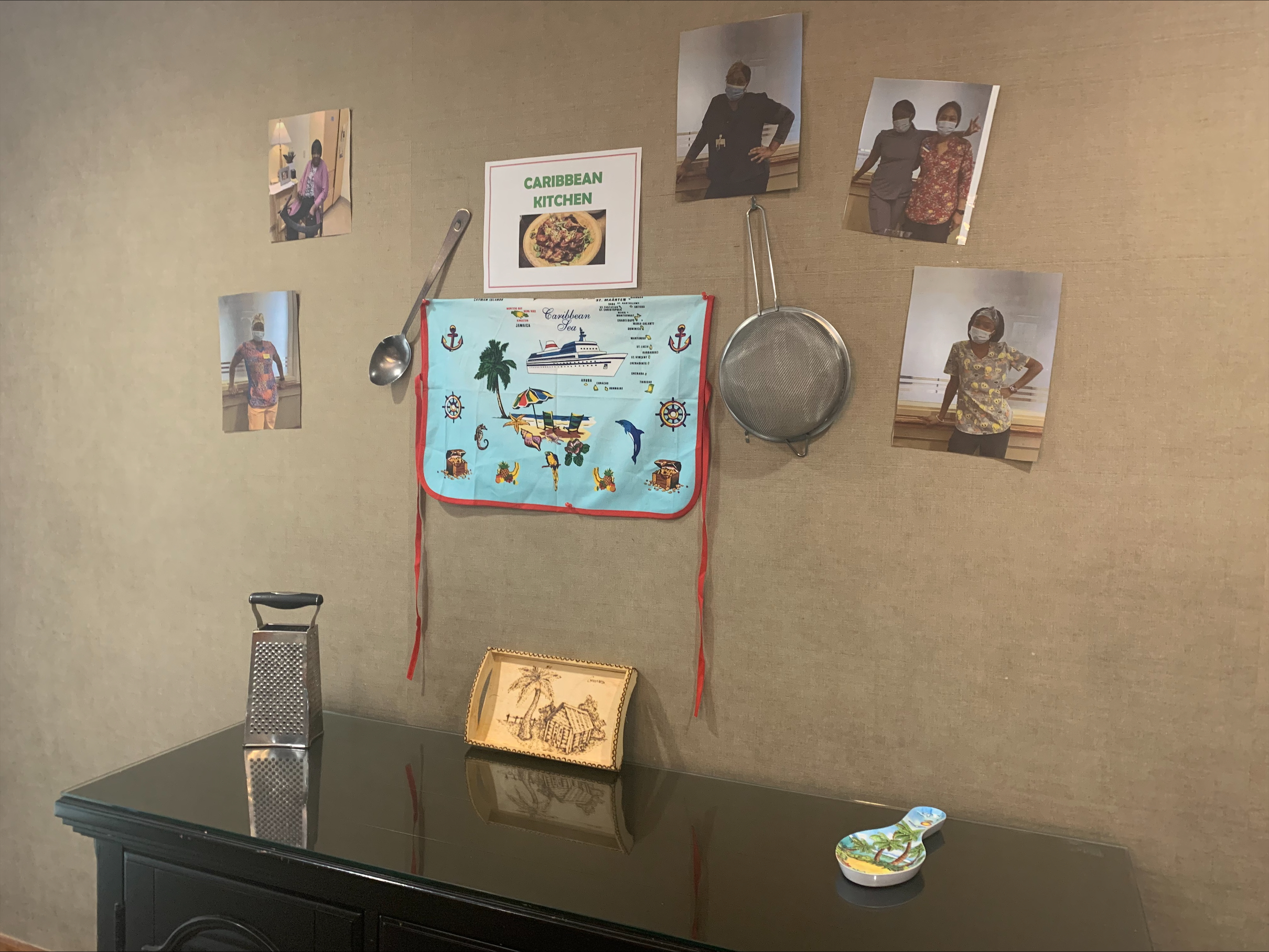 A wall display features a “Caribbean Kitchen” sign, photos of people, kitchen utensils, a decorative map, and a grater on a black table.