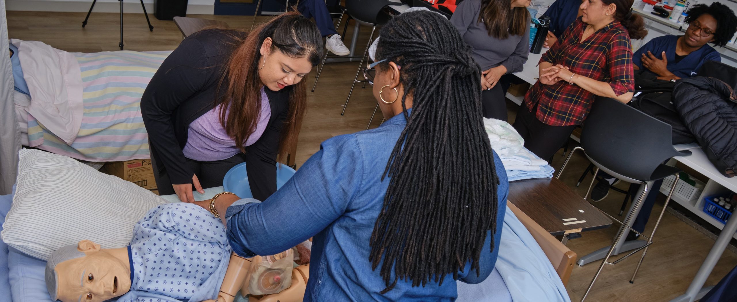 A group of people practice medical procedures on a hospital bed mannequin in a classroom setting.