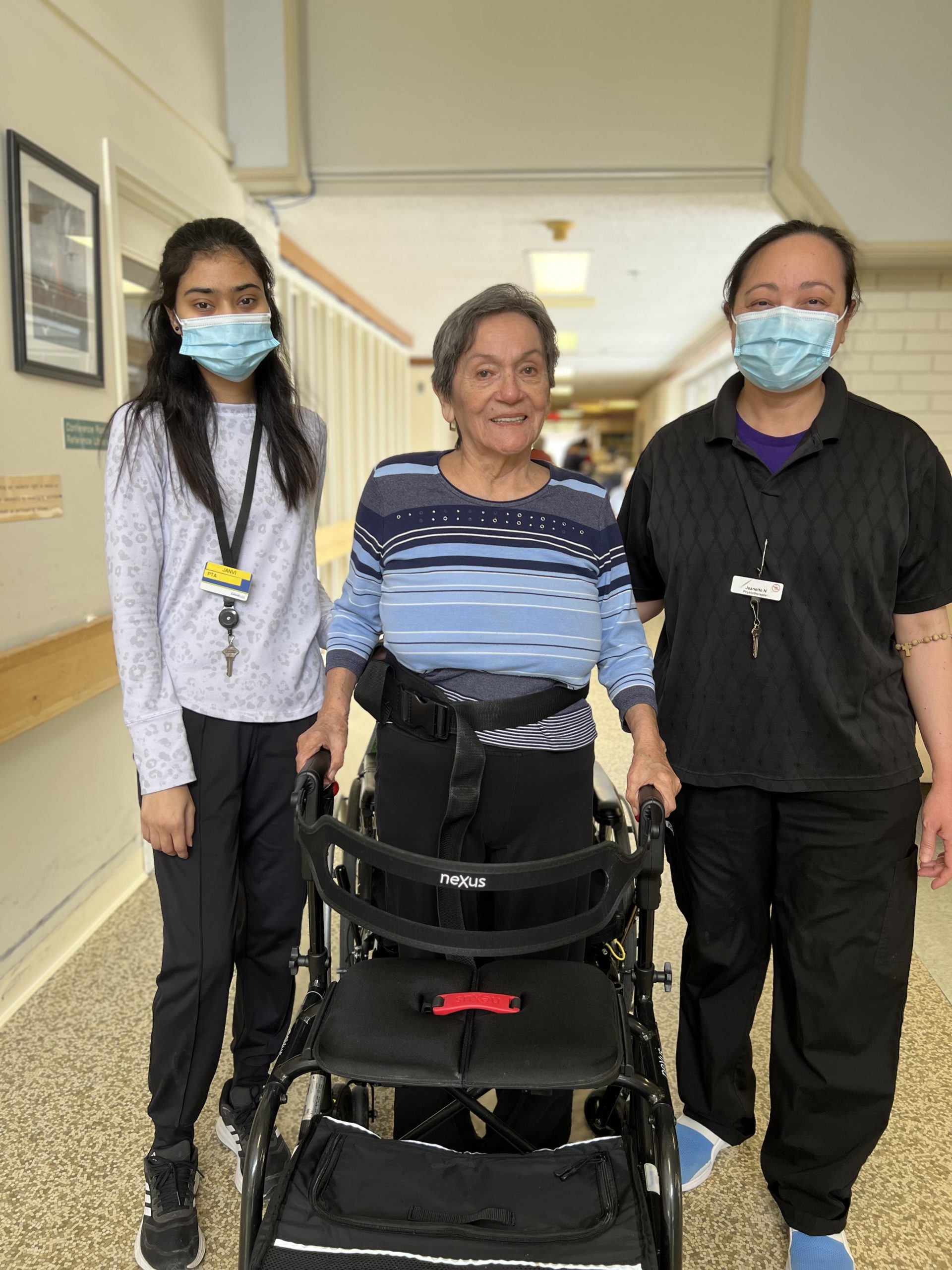 Three women, two in medical scrubs and masks, stand beside an elderly woman using a walker in a hallway.