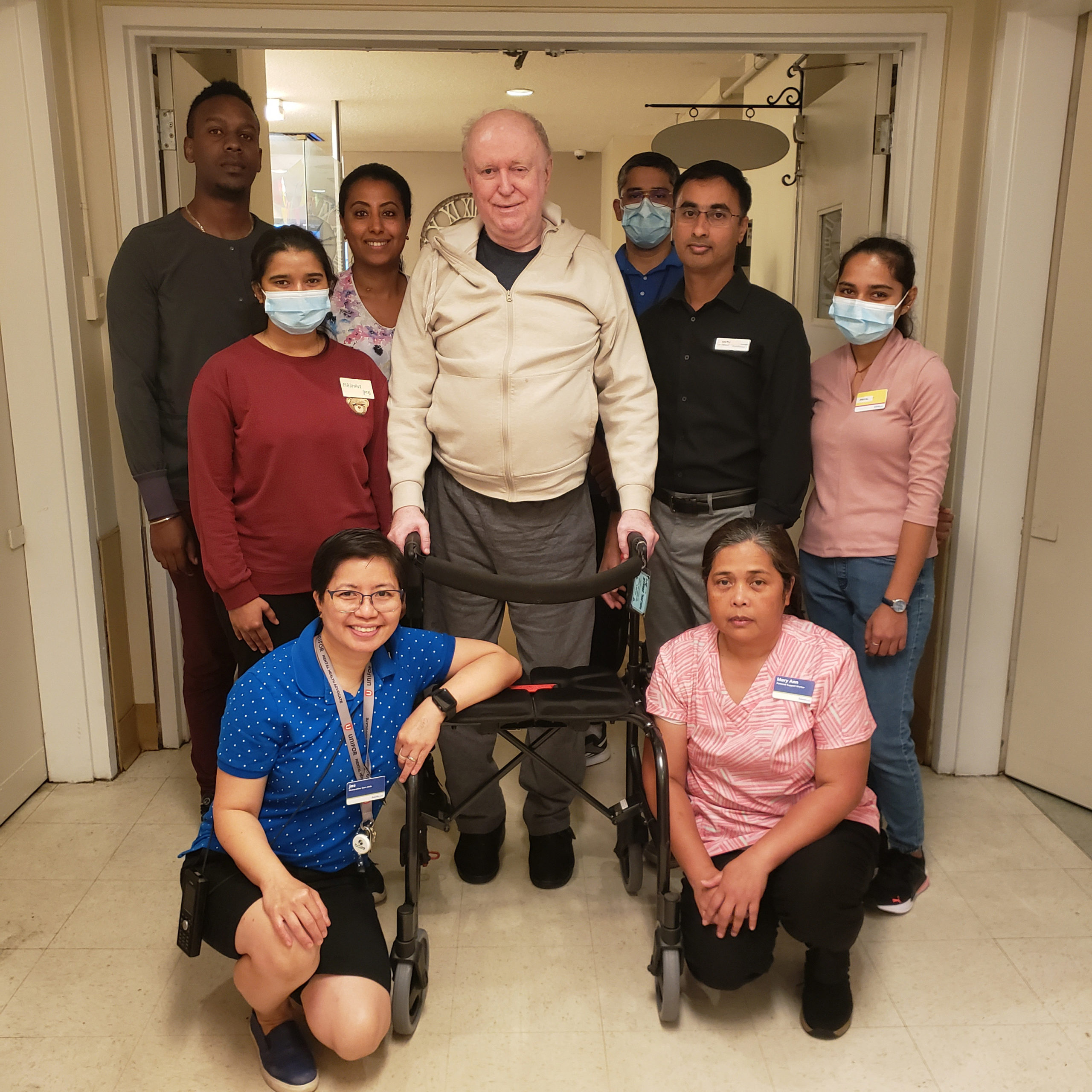 A group of eight healthcare workers pose around an older man with a walker, standing in a hallway of a care facility.