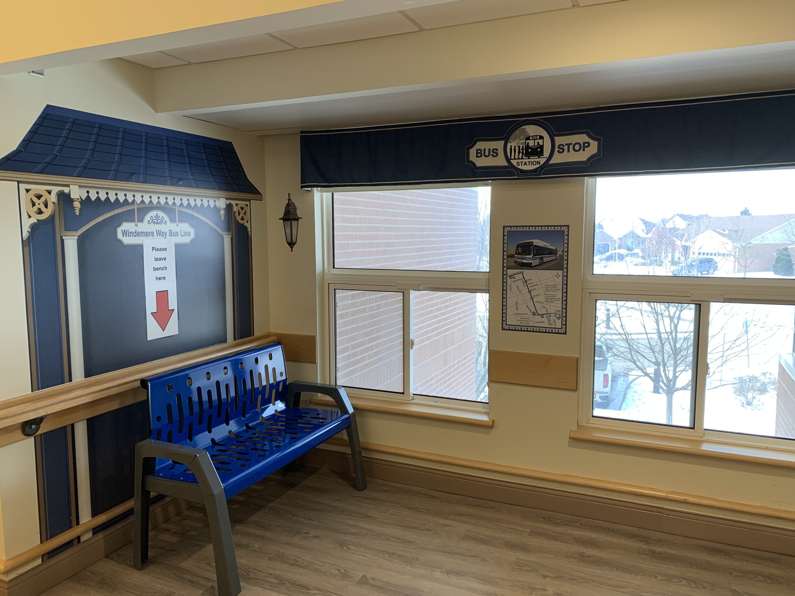 A blue bench sits by windows next to a decorative wall designed to resemble a bus stop, with a sign labeled "Wilderness Way Bus Line" and schedule details.