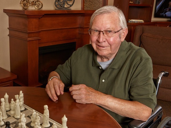 An older man in a green polo shirt sits in a wheelchair at a table with a chessboard in a warmly lit living room.