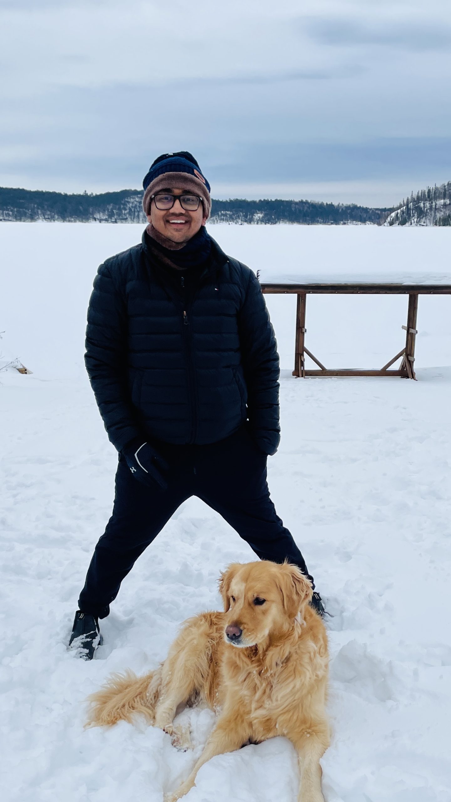 A man stands on snow wearing winter clothes and glasses, smiling, with a golden retriever lying at his feet; a frozen lake and snow-covered hills are in the background.