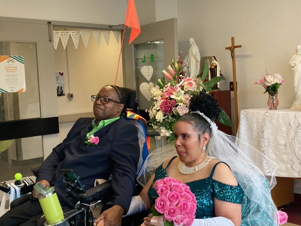 A couple dressed in formal attire, with the woman in a wedding dress holding pink roses and the man in a suit seated in a wheelchair, pose together in a decorated indoor setting.