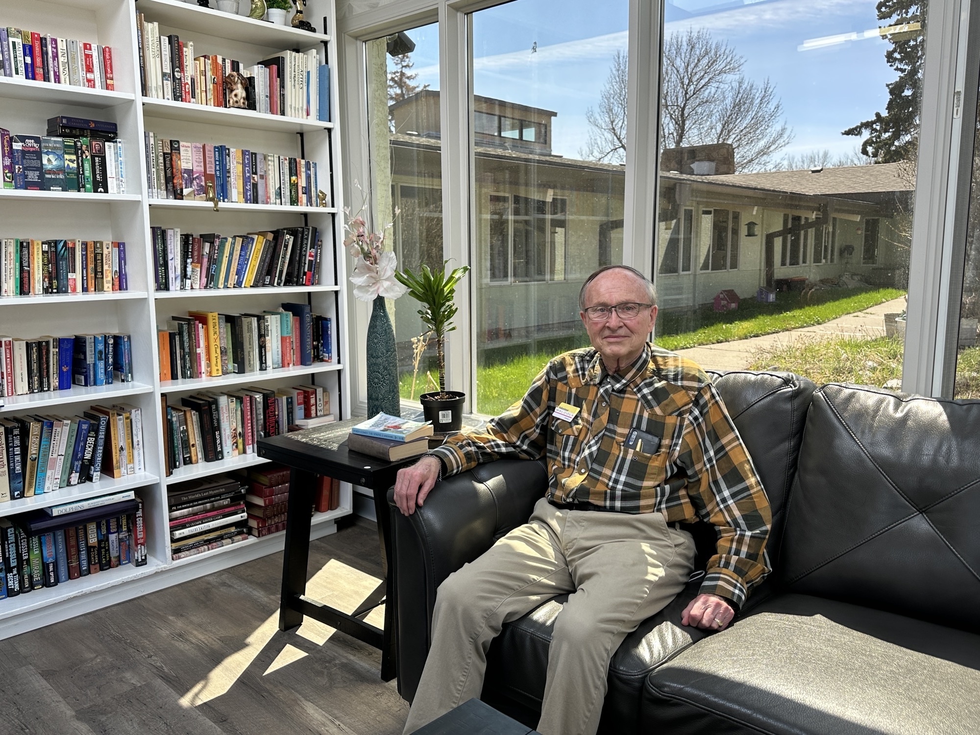 An older man sits on a black leather sofa in a sunlit room with large windows, next to a bookshelf filled with books and a small table with a plant.