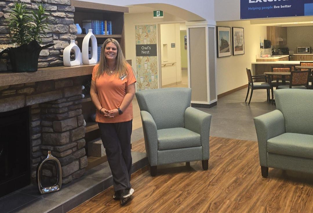 A woman in scrubs stands by a fireplace in a community room with two armchairs, a bookshelf, and a view into a kitchen area in the background.