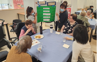 Six people sit around a table while two others stand and present information on a green board in a communal indoor setting.