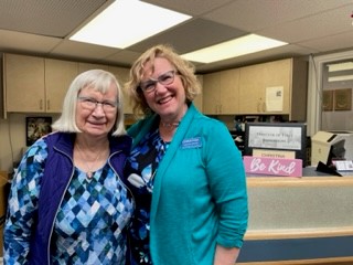 Two smiling women stand together in an office setting, both wearing glasses and blue clothing. A sign in the background reads "Be Kind.