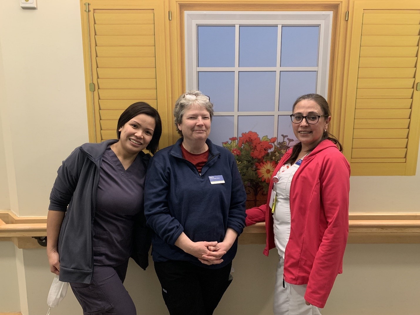 Three women in work uniforms stand indoors in front of a wall with a painted window and yellow shutters, posing and smiling at the camera.