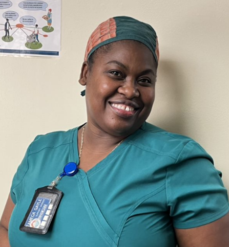 A person in teal medical scrubs and a head covering smiles at the camera. They wear an ID badge and are standing in front of a beige wall with a poster visible in the background.