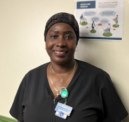 A woman in medical scrubs and a head covering stands indoors, smiling at the camera. She wears a badge and lanyard, with a health care poster visible on the wall behind her.