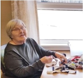 An elderly woman sits at a table near a window, sorting a pile of assorted socks and clothing items.