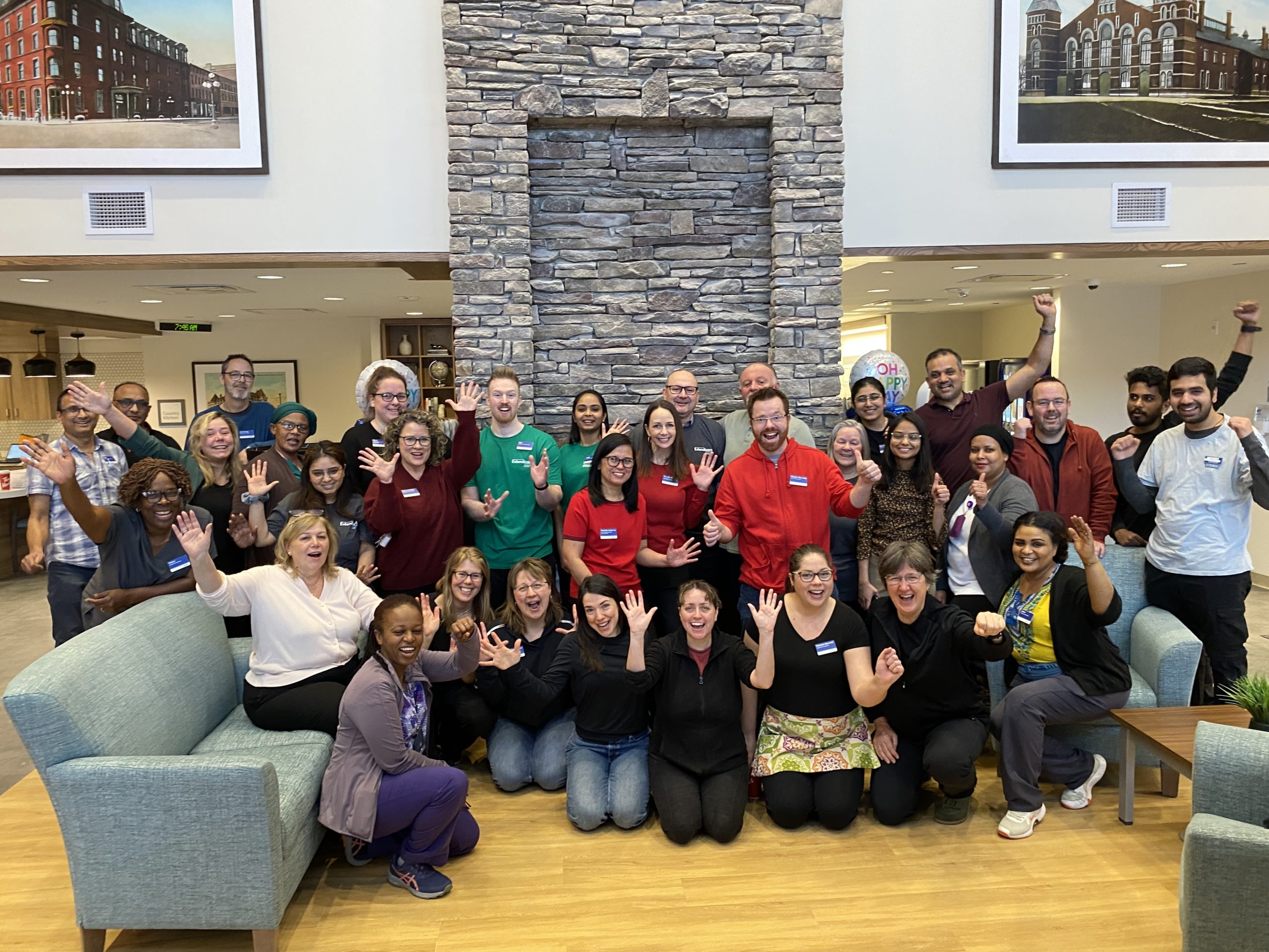 A group of adults poses indoors in front of a stone fireplace, smiling and waving at the camera. Some are standing, while others are seated on couches or kneeling on the floor.