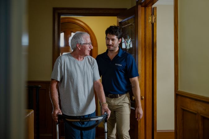 An older man using a walker is assisted by a younger man in a blue polo shirt as they walk through a hallway.