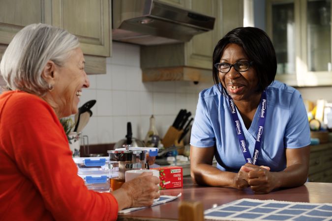 An elderly woman and a caregiver in scrubs talk and smile at a kitchen table with tea and medication organizers on the counter.
